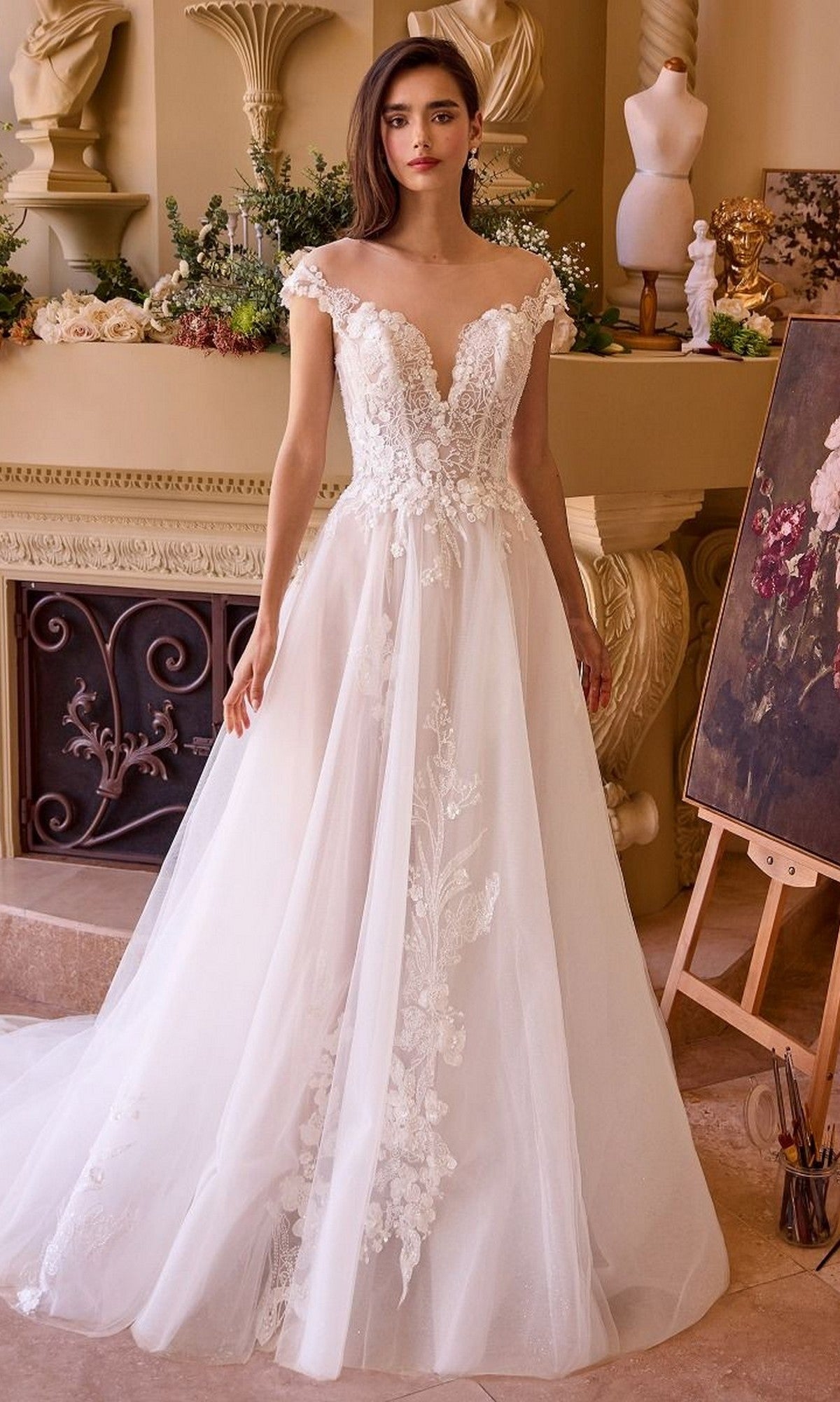 A woman wearing the Andrea and Leo WL038 long wedding dress with lace details stands indoors near classic decor and an easel displaying a painting.